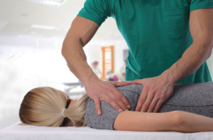 female dancer with blonde hair in ponytail laying face down on the physical therapy table, male physical therapist is working on patient's shoulder