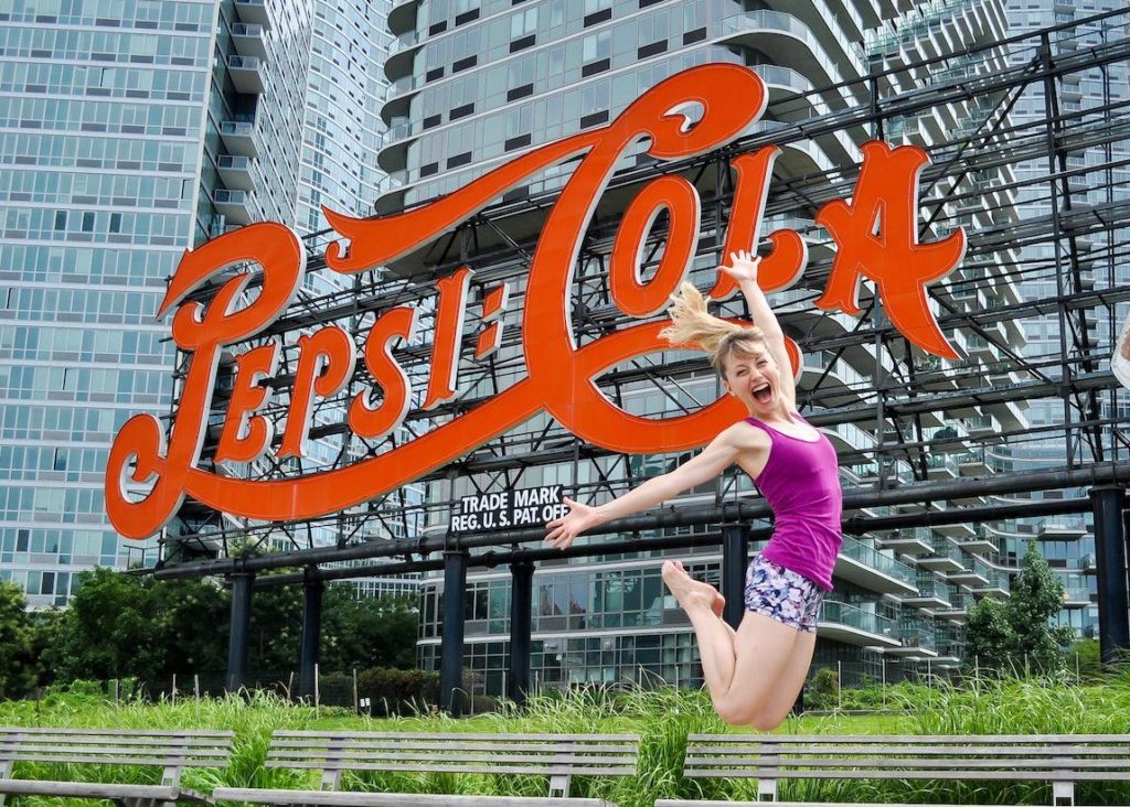 Move With HPA For Dancers founder Heidi Friese jumping for joy in front of Pepsi sign In NYC